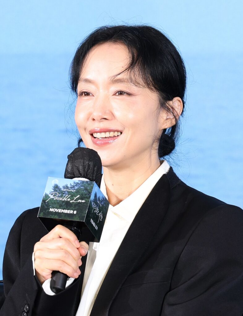 Jeon Do-yeon holding a microphone, wearing a black blazer and white shirt against a blue backdrop.